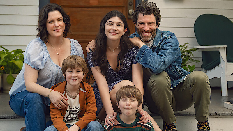 A family of five, including two adults, a teen girl, and two young boys, sits together smiling on the front porch steps of a house with white siding, plants, and a wooden door, suggesting a warm, casual outdoor setting.