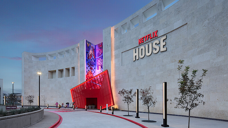 Modern building with large NETFLIX HOUSE sign, curved stone facade, red entrance, colorful mural, small trees, people approaching at dusk under a clear sky.