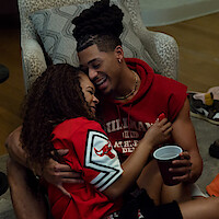 Two young adults laughing and hugging on the floor of a cozy living room, wearing casual sports jerseys. One holds a red plastic cup, and they appear happy and relaxed, enjoying each other’s company in a comfortable home setting.