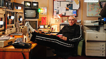 Vinnie Jones in a tracksuit relaxes with feet on desk in a cluttered control room, surrounded by monitors, electronics, bulletin board, and office equipment, holding a red cup.