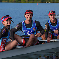 Three smiling rowers in team uniforms sit in a racing shell on a calm lake, wearing hats and sunglasses, with trees and water in the background, ready for a rowing competition or practice.