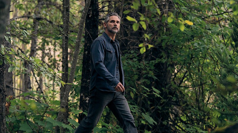A man with gray hair and a beard walks through a dense, green forest wearing a dark jacket and jeans. The environment is lush with foliage, trees, and natural light filtering through the leaves.