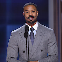 Michael B. Jordan in a gray suit speaking on stage at a formal event, standing behind a microphone with an award statue nearby, under professional lighting with a blurred blue and brown background.