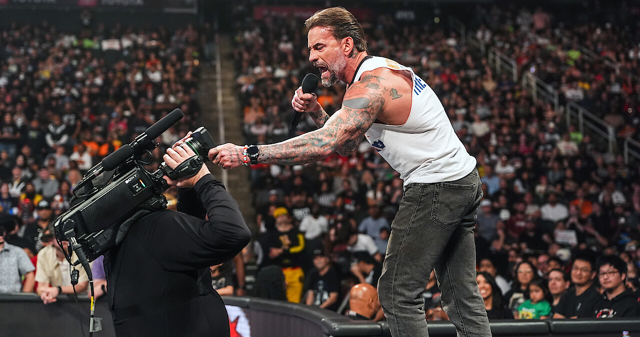 Tattooed man in a sleeveless shirt holding a microphone shouts into a camera at a crowded indoor arena, with audience watching intently from the stands and ringside area.