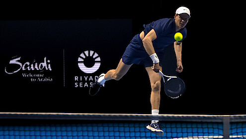 Male tennis player in navy outfit serves the ball on an indoor court during a match, with Saudi tourism and Riyadh Season logos visible in the background.
