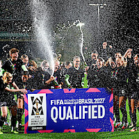 Women's soccer team celebrating qualification for FIFA Women’s World Cup 2027 in Brasil, spraying champagne on a lit outdoor field at night with a large "QUALIFIED" banner.