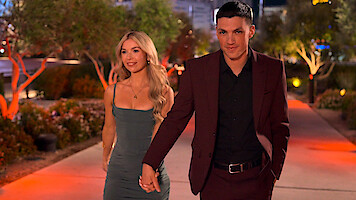 A man in a burgundy suit and a woman in a gray dress are holding hands while walking outdoors at night on a path lined with trees and decorative lights, suggesting a formal or romantic occasion.