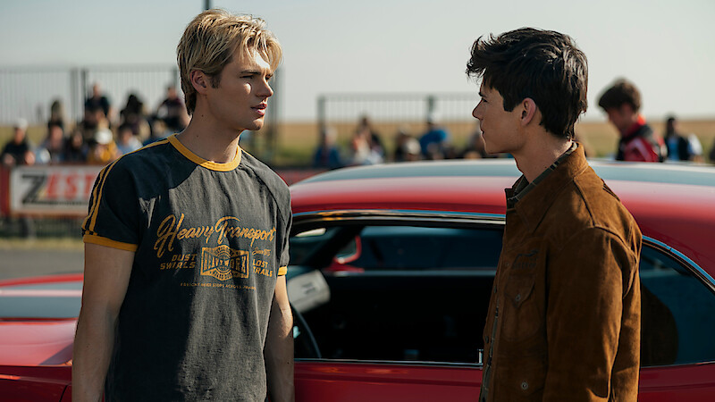 Two young men stand facing each other next to a red car at an outdoor event, with spectators and a fence visible in the background on a sunny day.