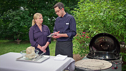 Two people in chef attire stand outdoors by a grill and table with a whole chicken in a bowl, discussing cooking preparations in a garden setting with trees and greenery in the background.