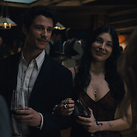 A man and woman dressed formally holding wine glasses, smiling at a social gathering in a warmly lit indoor setting with other people and pendant lights in the background.