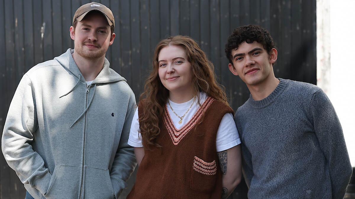 Three young adults standing side by side outdoors in front of a dark wooden wall, smiling at the camera. Casual clothing, natural lighting, and a relaxed atmosphere.