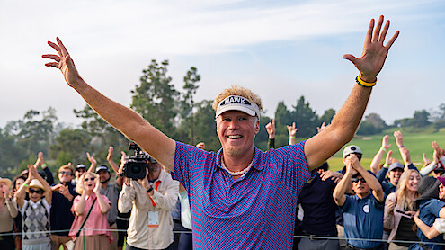 Smiling golfer with raised arms celebrates on a golf course, surrounded by cheering spectators and trees in the background, under a partly cloudy sky.