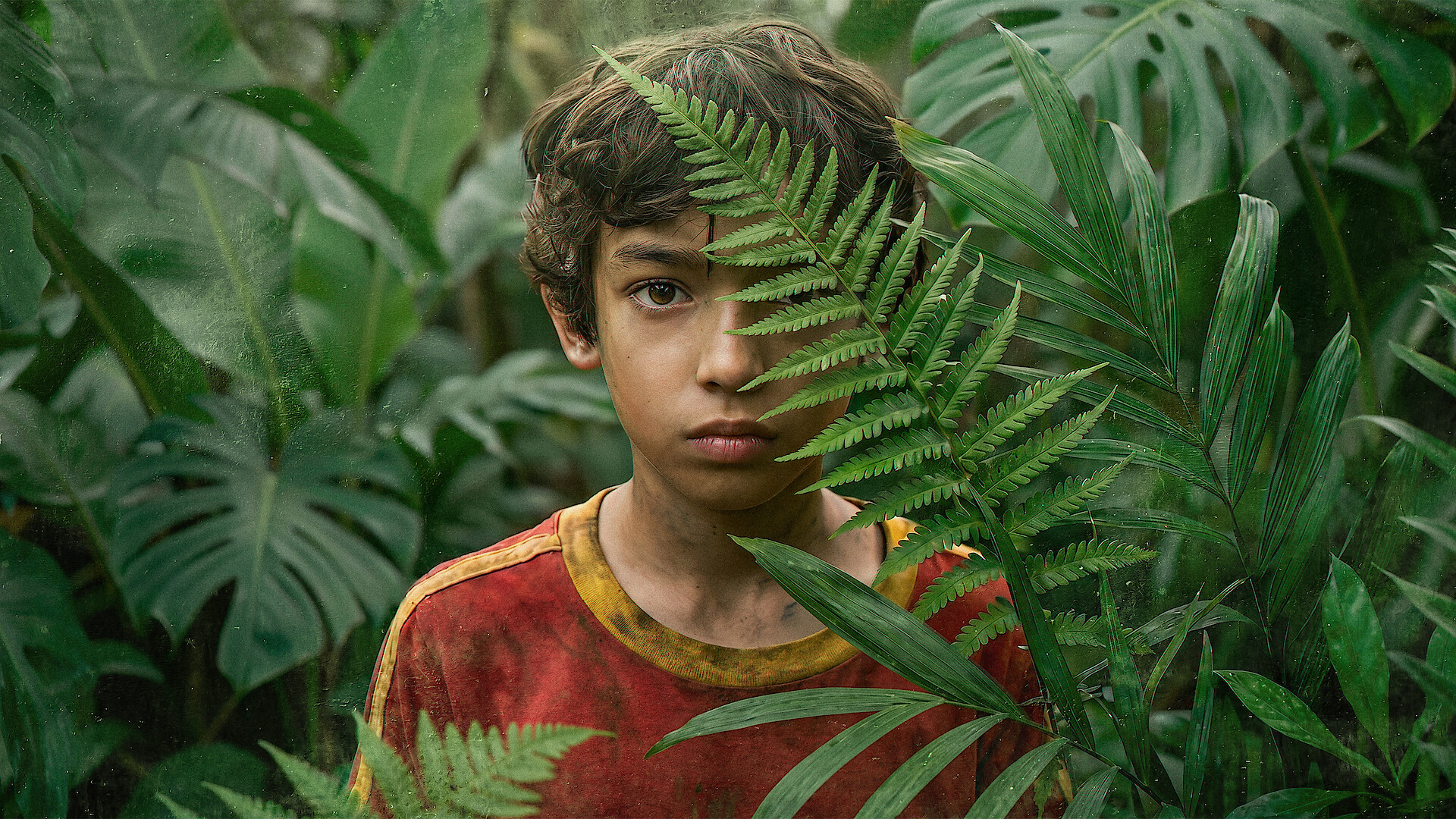 A young boy wearing a red shirt stands in a dense jungle surrounded by green tropical plants, partially obscured by a fern leaf, looking directly at the camera.