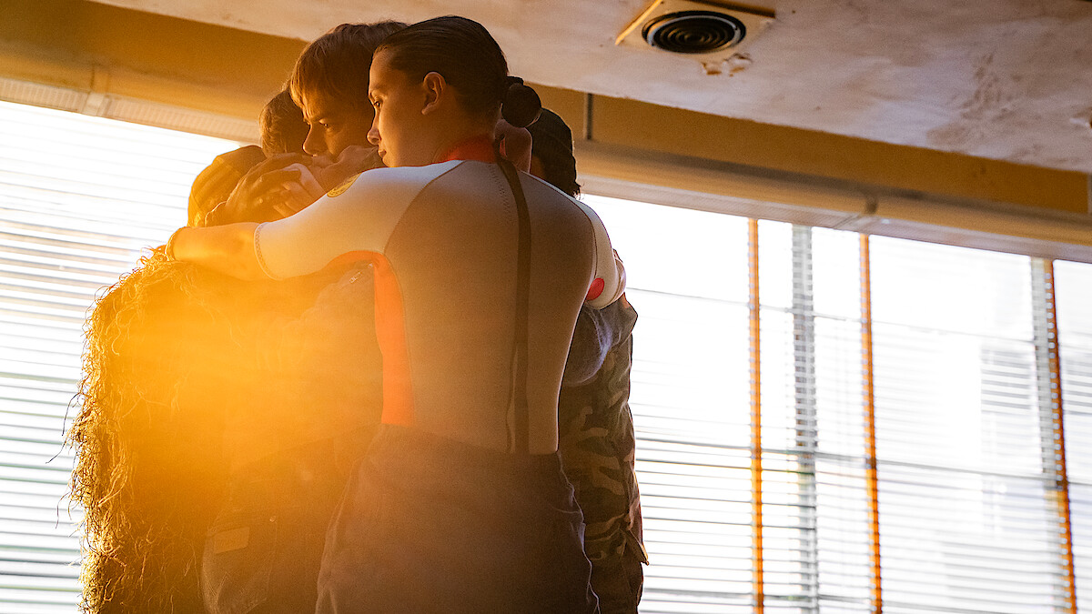 A small group huddles in a tight embrace, backlit by warm sunlight streaming through large window blinds.
