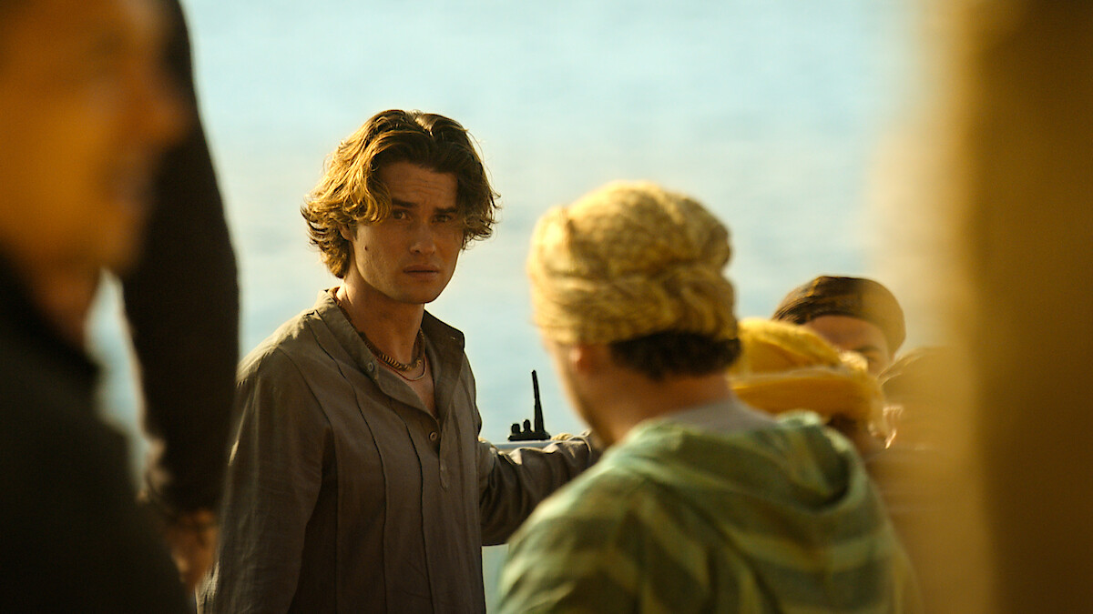 A young man with wavy hair stands outdoors near water, holding a walkie-talkie, looking toward the camera, surrounded by people dressed in casual clothing under warm sunlight.