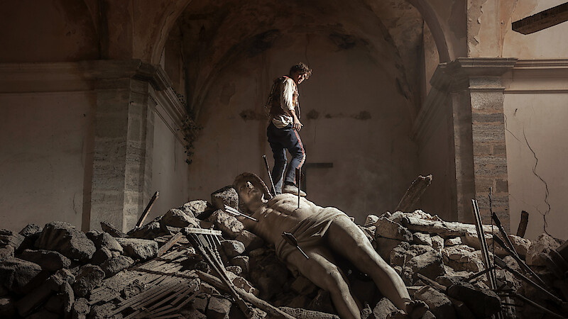 A person standing on rubble inside a damaged, old stone building, with a large fallen classical statue lying among the debris under an arched ceiling, evoking a sense of destruction and loss.
