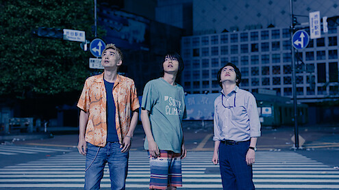 Three young men stand on a city crosswalk at night, looking up with curious or concerned expressions. The urban street is quiet and dimly lit, surrounded by buildings and street signs.