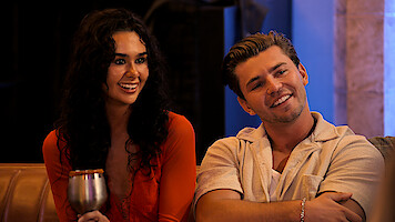Two young adults sitting on a couch indoors, smiling and looking toward someone off camera. The woman holds a drink, both appear relaxed. Warm lighting suggests a social, casual atmosphere.