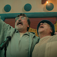Two older men singing passionately together into a microphone on stage, with festive decorations and warm lighting in the background.