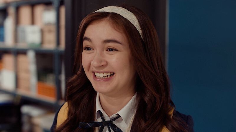 Smiling young woman with a headband and school uniform in an indoor setting, standing near blue wall and bookshelves.