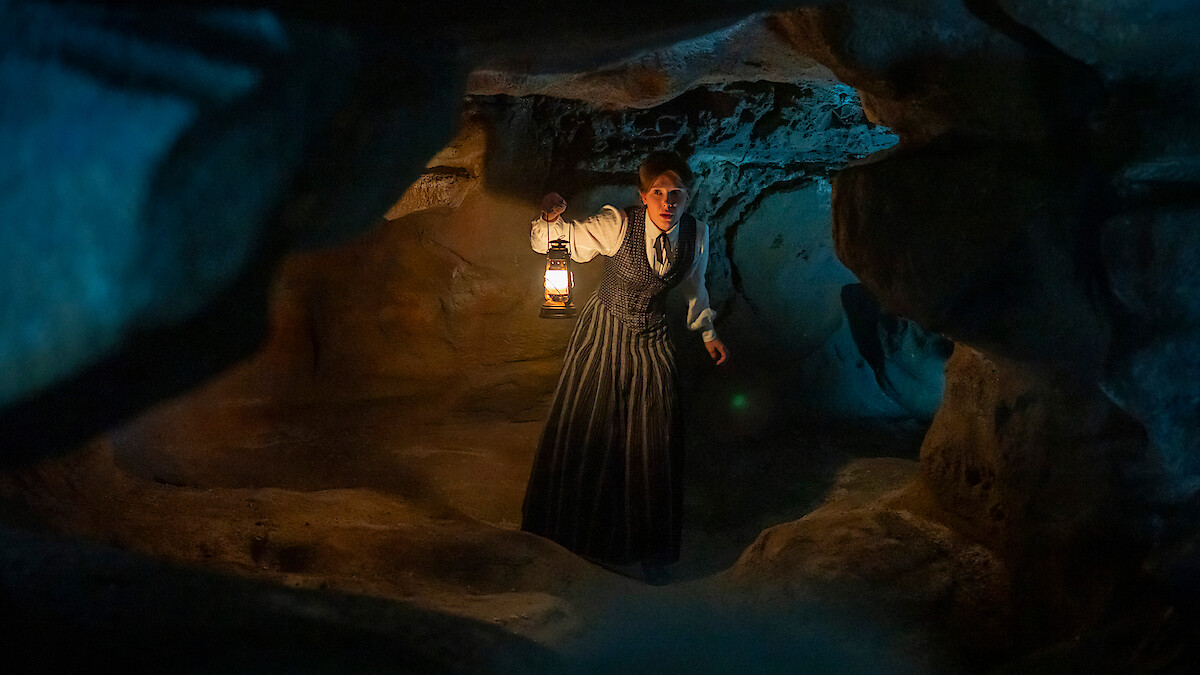 Woman in historical clothing holding a lantern exploring a dimly lit rocky cave, surrounded by shadows and uneven cave walls, with a mysterious blue light in the background.