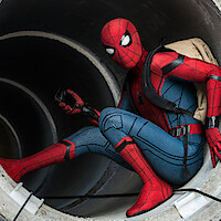 A person in a red and blue superhero costume crouches inside a large concrete pipe, holding a device and wearing a beige backpack in an urban, industrial setting.