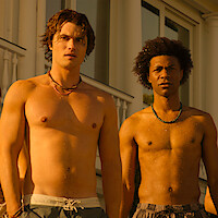 Two young men standing shirtless and wet outdoors, wearing swim trunks and necklaces, in front of a white building with columns and large windows, illuminated by golden sunlight.