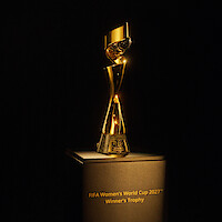 Golden FIFA Women’s World Cup 2027 Winner’s Trophy displayed on a pedestal, showcased in a dark environment with focused spotlight highlighting its shiny, elegant design and engraved base.