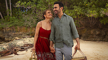A happy couple holding hands and carrying beach bags, walking on a sandy beach with trees and rocks in the background, enjoying a pleasant outdoor day together.