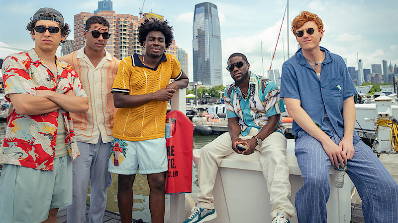Five young men in casual summer outfits stand by a marina with boats, modern city buildings, and a waterfront skyline in the background, suggesting a relaxed and urban environment.