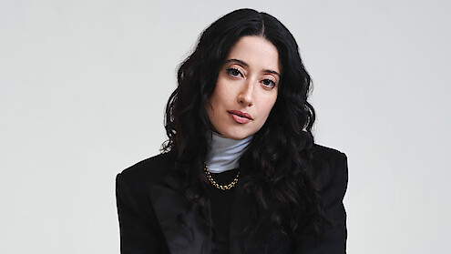 Taylor Ortega photographed in front of a white backdrop wearing a black jacket with strong shoulders, white turtle neck layered under a black shirt, and a gold chain with her dark curly hair cascading around her face.