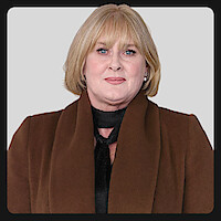 A woman with short blonde hair wearing a brown coat and black scarf poses against a plain light gray background in a studio or professional setting.