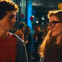 A man and woman talking at a lively indoor party with colorful balloons and people socializing in the background, warm lighting.
