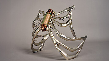 Futuristic spider-like sculpture with metallic legs and a copper coil body, set against a gray background. The mood is mechanical and surreal, evoking curiosity and intrigue.