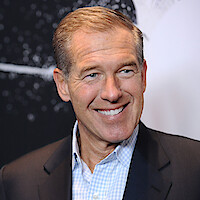 Man in a suit smiling in front of a dark, abstract background with white speckles and lines, possibly at an event or exhibition.