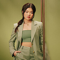 Miyako, a woman in a light green suit and crop top stands confidently against a matching green background, with one hand in her pocket, her hair styled in a loose side braid, conveying a modern, stylish atmosphere.