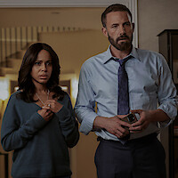A concerned woman and a serious man in business attire stand indoors, possibly in a living room with warm lighting and a staircase in the background, suggesting a tense or emotional moment.