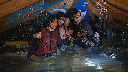 Four people waist-deep in floodwater in an attic, huddled together, appearing worried and alert, with one person holding a weapon, water pouring from ceiling, scene is tense and dramatic.
