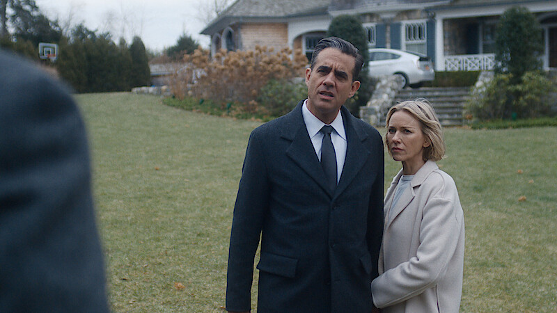 Man in dark coat and woman in light coat stand together on a lawn, looking concerned, in front of a large house with stone steps and autumn landscaping.