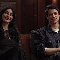 Camila Morrone and Adam DiMarco sitting on a couch in a cozy, warmly lit room with wooden walls, smiling and relaxed.