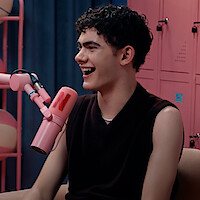 Joe Locke smiling and speaking into a pink microphone with a red 'N' in a colorful podcast studio featuring pink lockers and a blue curtain.