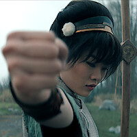 Miyako as Toph dressed in ancient-style clothing and headband poses with closed fist outdoors, with a wooden staff in the background and blurred grassy landscape.