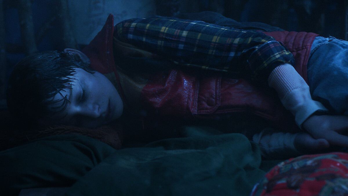 A young boy in a plaid shirt and red vest lies curled up on his side in a dimly lit setting, appearing wet, alone, and cold.