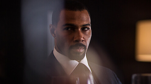 A man in a suit sits in a dimly lit room with a serious expression, blurred glass and a lamp in the background, creating a dramatic, tense atmosphere.