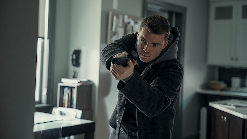 A man holding a gun in a ready position stands alert in a modern, dimly lit kitchen or office environment, appearing tense and focused as he aims at something off-frame.