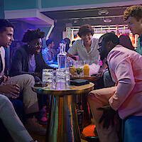 Five young men sit around a table in a lively nightclub, talking and laughing together. The table holds drinks, ice, and glasses, with colorful lighting and other people mingling in the background.