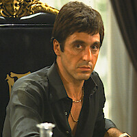 Man with serious expression sits at an ornate black chair with gold details, wearing a dark shirt and gold chain, indoors near a table in a formal setting with rich decor.