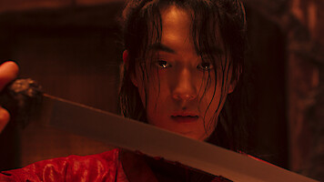 Person with long hair and wet face in red robe holding a sword, intense expression, standing indoors in dim, dramatic lighting with a blurred background.