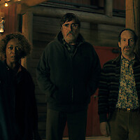 Three concerned adults stand together at a dimly lit wooden cabin, illuminated by warm light, looking ahead with tense expressions.