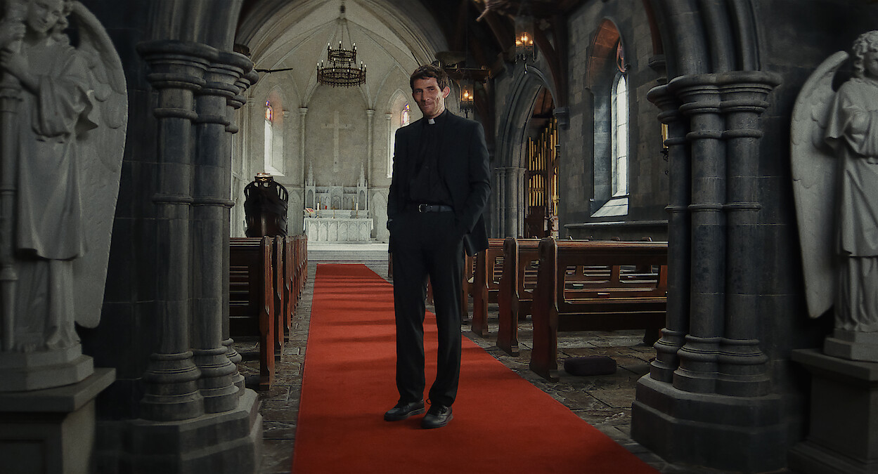 A priest stands on a red carpet in the middle aisle of a dimly lit stone church, flanked by statues, with pews on either side and an altar in the background, creating a solemn and contemplative mood.